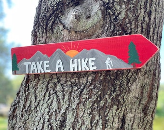 May include: A red wooden arrow-shaped sign with the words "TAKE A HIKE" in white. The sign features a mountain range, a rising sun, and a hiker silhouette. Green trees are painted on each end of the sign.