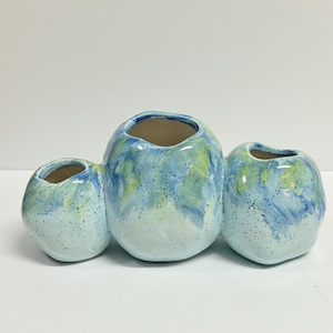 May include: A ceramic vase with three connected, rounded openings. The vase is painted with a blend of blue, green, and white hues, speckled with darker dots. The interior of each opening is a light brown color.