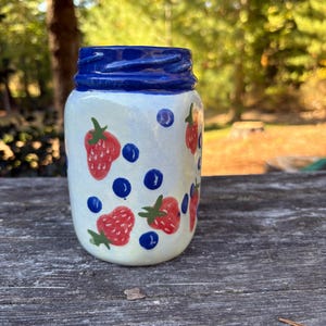 May include: A ceramic jar with a blue rim and a white body decorated with red strawberries and blue blueberries. The jar is cylindrical and appears to be hand-painted. The background is blurred, suggesting an outdoor setting.