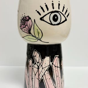 May include: A ceramic cup with a cream-colored top and a black base. The top features a black-outlined eye and a pink flower. The base is decorated with pink crystal formations. A unique, artistic piece.