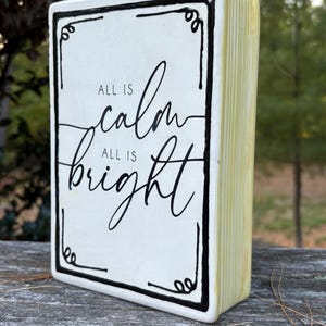 May include: A ceramic book-shaped decoration with the words "ALL IS calm ALL IS bright" in black script. The front is white with a black border and decorative corner elements. The sides are a pale yellow, giving the appearance of book pages.