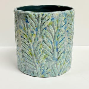 May include: A cylindrical ceramic planter with a textured leaf pattern in shades of blue, green, and yellow. The interior is a dark teal color. The planter is a decorative item for indoor plants.