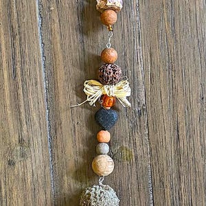 Könnte beinhalten: Handgefertigter Hängeornament mit verschiedenen Naturelementen. Es enthält Perlen, Steine und Filzelemente in Grün, Braun und Orange. Der Ornament ist an einem Ast aus Holz aufgehängt.