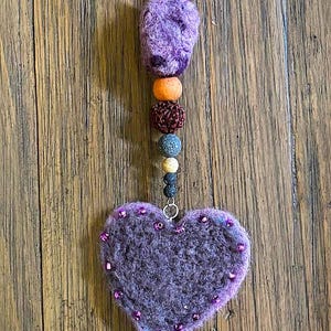 May include: A handmade wall hanging with a purple felt heart, wooden beads, and natural materials. The decorative item includes a beaded top and a heart-shaped felt design, with a rustic aesthetic.