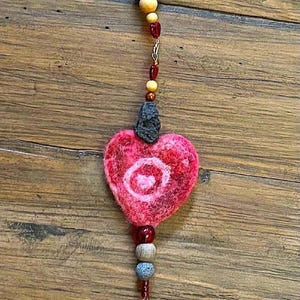 May include: A handcrafted wall hanging featuring a variety of natural elements. It includes a felt heart, wooden beads, and a decorative wooden branch. The piece is adorned with red and orange accents, creating a rustic aesthetic.