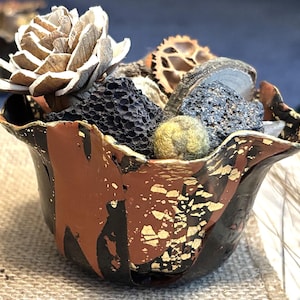 May include: A decorative bowl with a black, brown, and gold exterior, filled with various natural elements. The bowl contains a large, light brown, flower-like object, along with other textured items in shades of grey, brown, and green.
