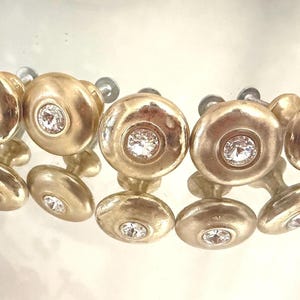 Puede incluir: Un conjunto de seis pomos dorados para armarios, cada uno con un cristal central brillante. Los pomos son redondos con una cara ligeramente abovedada y una pequeña base ovalada. Reflejos de los pomos en la superficie.