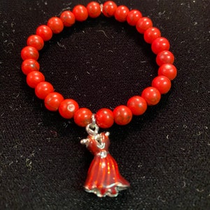 May include: A red beaded bracelet with a red dress charm. The beads are small and round, and the charm is detailed with a silver outline.