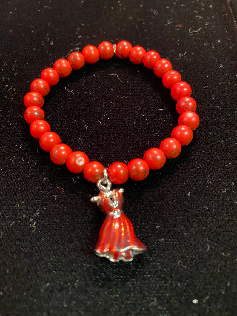 May include: A red beaded bracelet with a red dress charm. The beads are small and round, and the charm is detailed with a silver outline.