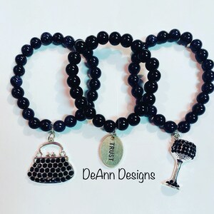 May include: Three black beaded bracelets with charms. The charms are a purse, a wine glass, and a tag that says "TRUST".
