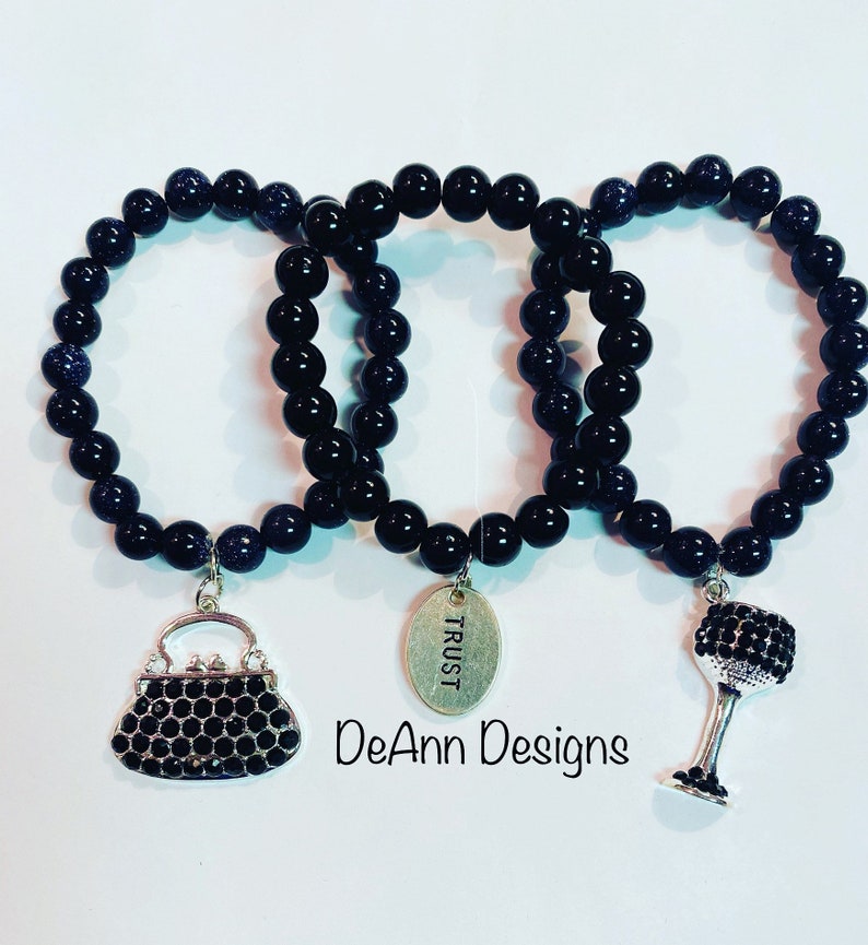 May include: Three black beaded bracelets with charms. The charms are a purse, a wine glass, and a tag that says "TRUST".