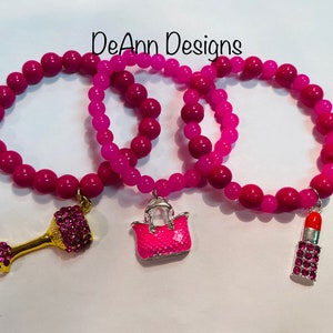 May include: Three pink beaded bracelets with charms. The charms are a wine glass, a handbag, and a lipstick.