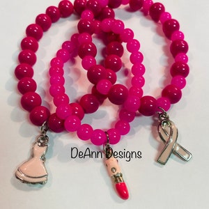 May include: Three pink beaded bracelets with charms. The charms are a pink lipstick, a white dress, and a pink ribbon. The bracelets are stacked on top of each other and the charms are hanging down. The text "DeAnn Designs" is visible in the image.