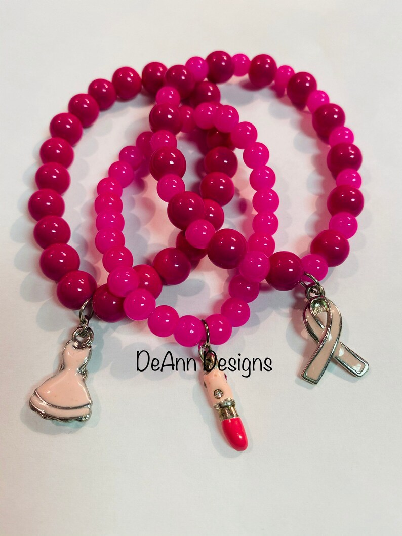 May include: Three pink beaded bracelets with charms. The charms are a pink lipstick, a white dress, and a pink ribbon. The bracelets are stacked on top of each other and the charms are hanging down. The text "DeAnn Designs" is visible in the image.