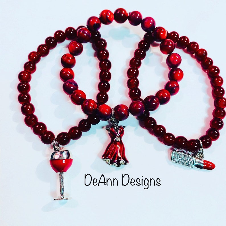 May include: Three red beaded bracelets with charms. The charms are a red dress, a wine glass, and a lipstick. The bracelets are stacked on top of each other. The text "DeAnn Designs" is visible below the bracelets.