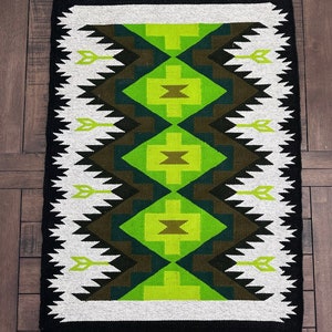 May include: A black and white rug with a geometric pattern in shades of green and brown. The rug has a fringed border.