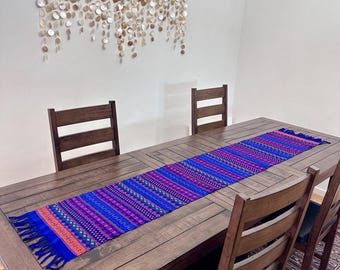 Op de afbeelding: Een paarse, blauwe en roze tafelkleed met franjes aan de uiteinden. De tafelkleed ligt op een houten tafel met vier stoelen. Een schelpenwanddecoratie hangt aan de muur achter de tafel.