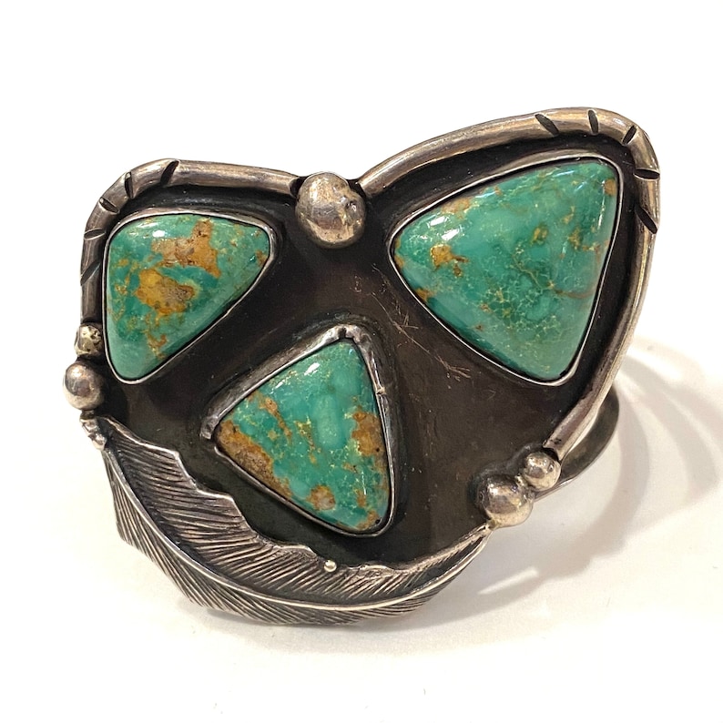 May include: A silver bracelet with three turquoise stones set in a triangular shape. The bracelet has a feather design on the bottom.