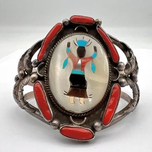 Puede incluir: Un brazalete de plata con una gran incrustación ovalada que representa a una mujer nativa americana con un vestido turquesa y rojo. La incrustación está rodeada de piedras de coral rojo y detalles plateados.