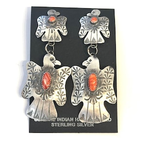 May include: A pair of sterling silver drop earrings featuring a two-tiered thunderbird design. Each bird is stamped with intricate patterns and set with an oval orange stone in the centre. The earrings are displayed on a black card.