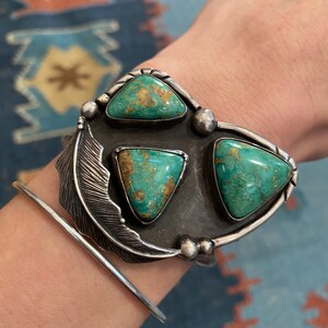 May include: A silver cuff bracelet with a feather design and three turquoise stones. The stones are triangular in shape and have a green and brown color.