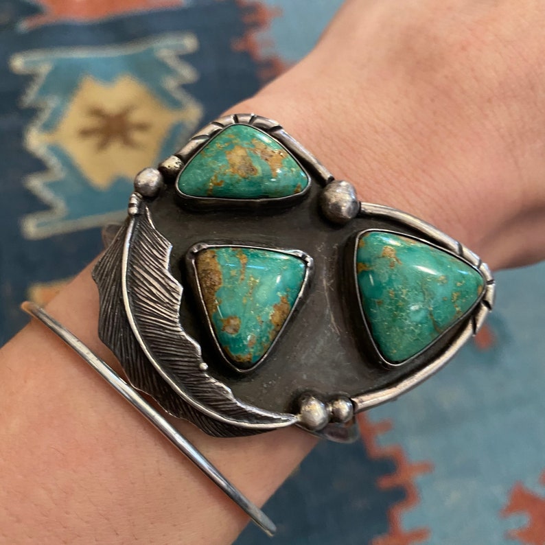May include: A silver cuff bracelet with a feather design and three turquoise stones. The stones are triangular in shape and have a green and brown color.