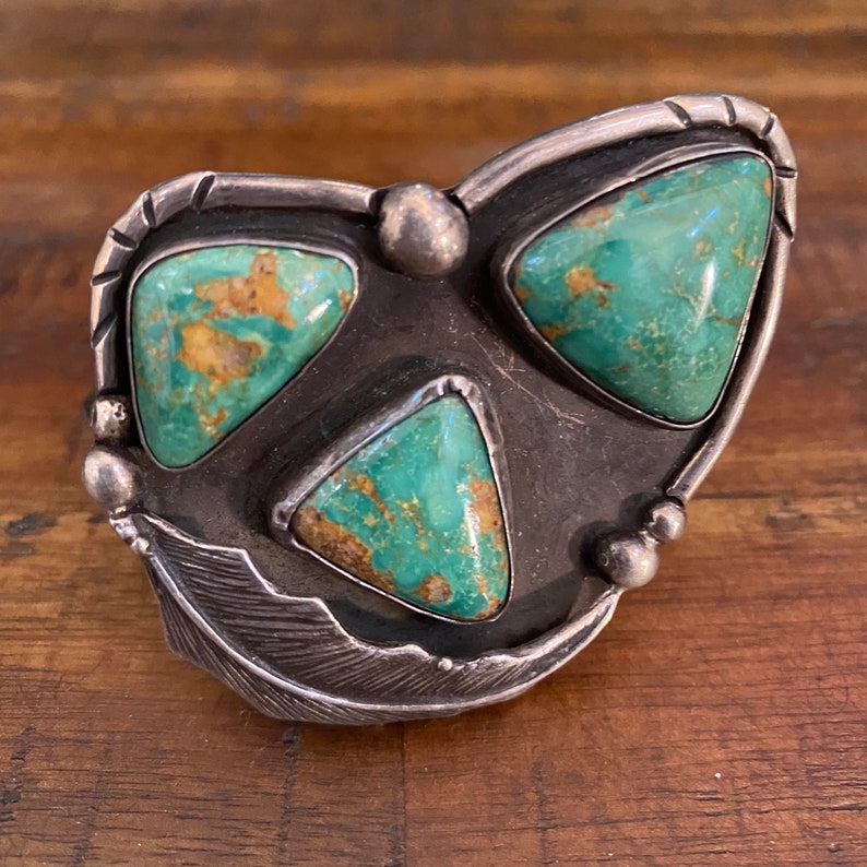 May include: A silver and turquoise Native American ring with a heart-shaped design and three turquoise stones. The ring has a feather design on the side.