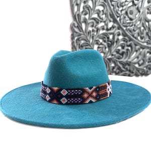 May include: Teal felt fedora hat with a colorful woven band. The band features geometric patterns in shades of pink, brown, white, and black.