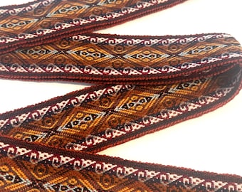 May include: A long, woven fabric strap featuring a repeating geometric diamond pattern in shades of deep red, burnt orange, brown, and light blue. The edges are finished with a solid dark red border, and the texture appears dense and durable.