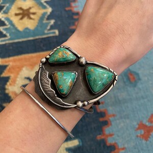 May include: A silver cuff bracelet with a turquoise stone design. The bracelet features three triangular turquoise stones set in a silver frame with a feather design.