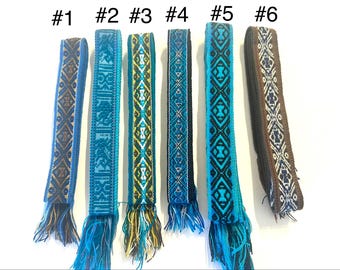 May include: Six woven fabric belts labelled #1 through #6, each featuring distinct geometric patterns. The belts display various colour combinations including blue, teal, yellow, black, and brown, all finished with fringed ends at the bottom.