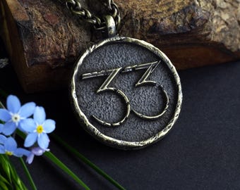May include: A round, bronze-toned pendant necklace with the number "33" in a stylized font. The pendant hangs from a matching chain. The background includes a piece of wood, blue flowers, and a dark surface.