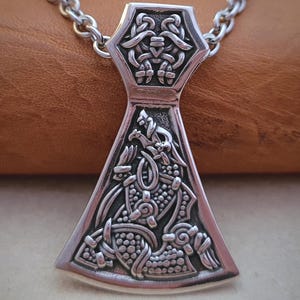 Puede incluir: Un collar con colgante plateado con un diseño detallado de cabeza de hacha vikinga. El colgante presenta patrones intrincados y está suspendido de una cadena a juego. La cabeza del hacha mide aproximadamente 5 cm de largo, mostrando un tema histórico o de fantasía.