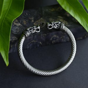 May include: A silver bracelet with a twisted rope design and two animal head accents. The bracelet is on a dark background.