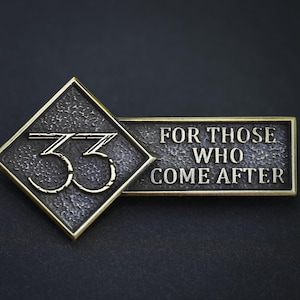 May include: A metallic pin featuring a diamond-shaped section with the number 33 in raised lettering and a rectangular section with the text FOR THOSE WHO COME AFTER. The piece has a dark, textured finish with gold-toned edges and raised details.