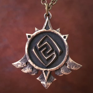 Op de afbeelding: Een bronzen ketting met een hanger met een gestileerd geometrisch ontwerp. De hanger is rond met een centraal symbool en een stervormig patroon rond de rand. De hanger is bevestigd aan een ketting.