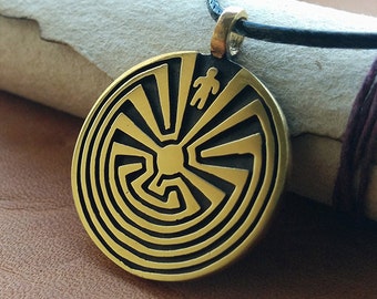 May include: A gold-colored pendant necklace with a black leather cord. The pendant features a circular design with a black and gold labyrinth pattern and a small figure in the center.