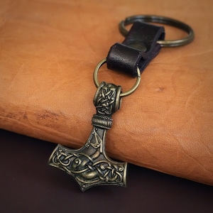May include: A bronze Mjolnir keychain with intricate knotwork details, attached to a brown leather strap with a silver key ring.