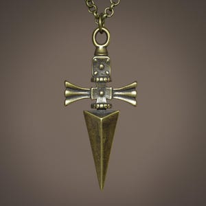 Pu&ograve; includere: Una collana con un ciondolo dorato a forma di pugnale stilizzato, con un design a forma di croce. Il pugnale ha una punta appuntita e un design dettagliato e decorato.