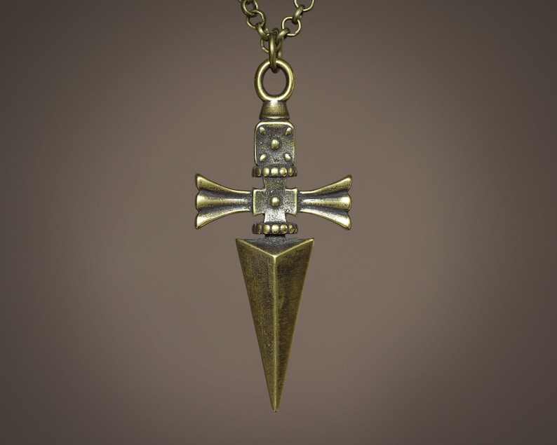 Pu&ograve; includere: Una collana con un ciondolo dorato a forma di pugnale stilizzato, con un design a forma di croce. Il pugnale ha una punta appuntita e un design dettagliato e decorato.