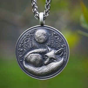 May include: A silver pendant necklace featuring a detailed, three-dimensional design of a fox curled up under a full moon in a forest setting.