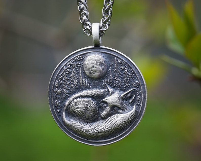 May include: A silver pendant necklace featuring a detailed, three-dimensional design of a fox curled up under a full moon in a forest setting.
