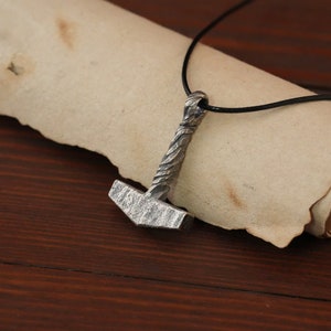 May include: A silver Mjolnir pendant necklace with a black leather cord. The pendant is detailed with a textured hammer head and a braided handle.