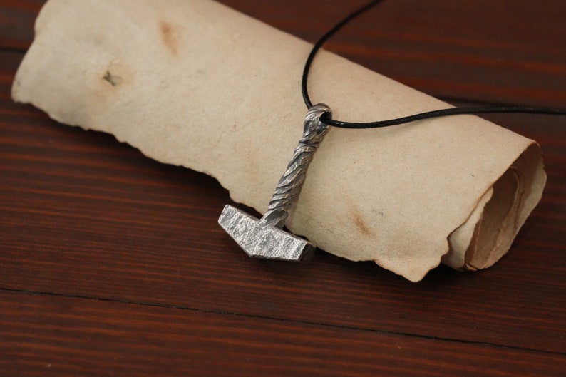 May include: A silver Mjolnir pendant necklace with a black leather cord. The pendant is detailed with a textured hammer head and a braided handle.