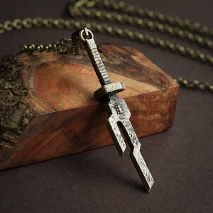 May include: A silver-toned sword pendant necklace with a chain. The sword has a detailed design and is hanging on a piece of wood.