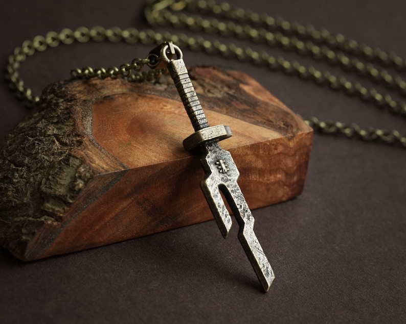 May include: A silver-toned sword pendant necklace with a chain. The sword has a detailed design and is hanging on a piece of wood.
