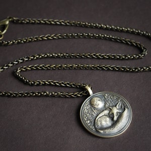 May include: A silver chain necklace with a round pendant featuring a detailed sculpture of a fox sleeping under a crescent moon.