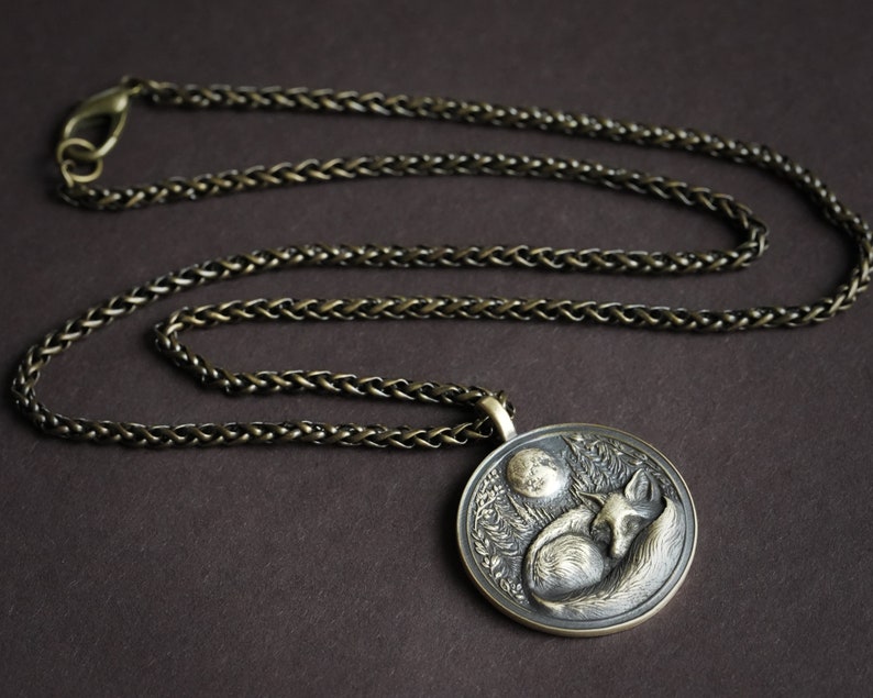May include: A silver chain necklace with a round pendant featuring a detailed sculpture of a fox sleeping under a crescent moon.