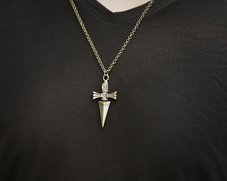 Pu&ograve; includere: Una collana dorata con un ciondolo a forma di spada. Il ciondolo presenta un design dettagliato con un motivo geometrico.