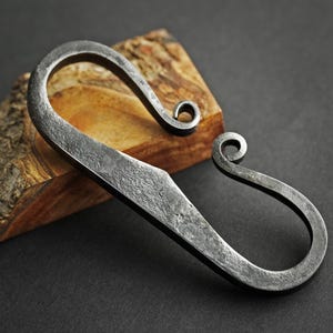 Puede incluir: Un iniciador de fuego de varilla de ferrocerio forjado a mano con un acabado de metal oscuro texturizado. La herramienta tiene un diseño curvo en forma de S con un extremo puntiagudo y un bucle rizado. Descansa sobre un trozo de madera con veta natural, sobre fondo oscuro.